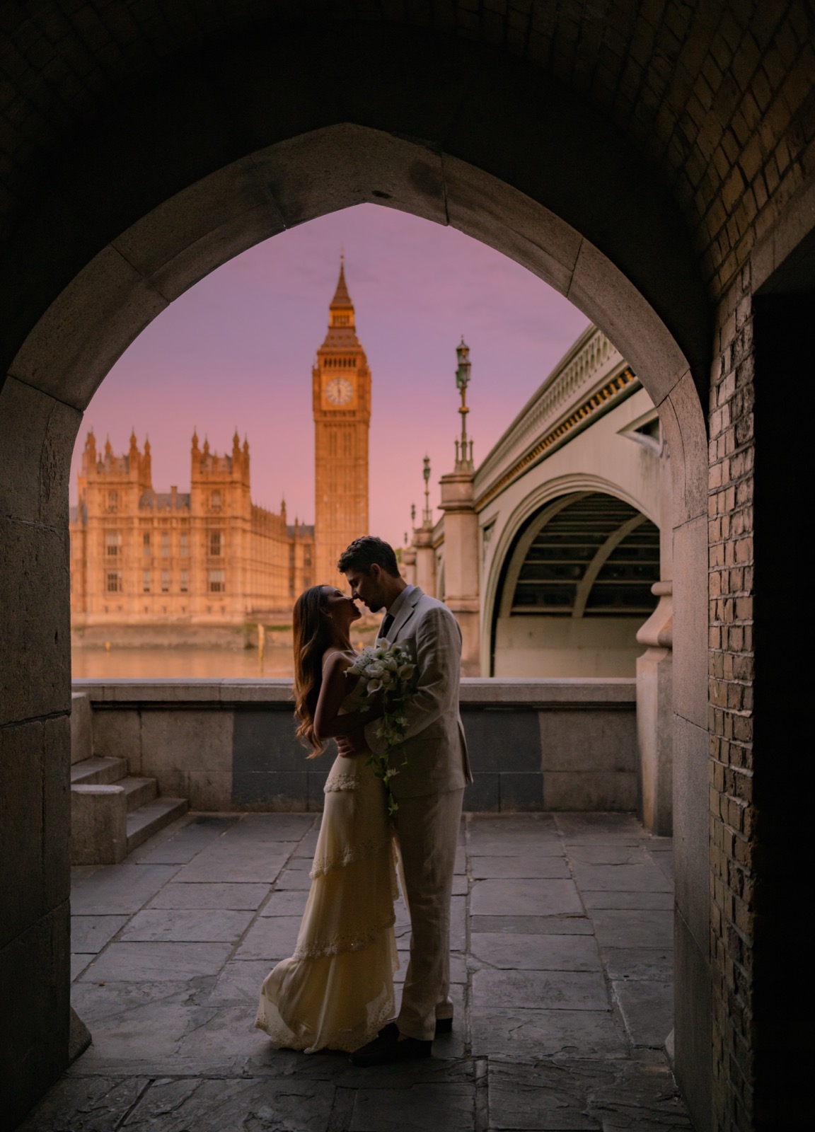 Rachel and Gustavo under a stone archway at Westminster, Big Ben and a pink sunset sky behind them